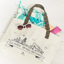 Cargar imagen en el visor de la galería, Bolsa de playa de tela beige con ilustración de Alicante y asas de cinta negra y beige, con toalla turquesa, gafas de sol y cremas en el interior.