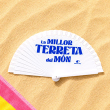 Cargar imagen en el visor de la galería, Abanico blanco de madera abierto con el texto azul “La millor terreta del món” sobre arena de playa de Alicante
